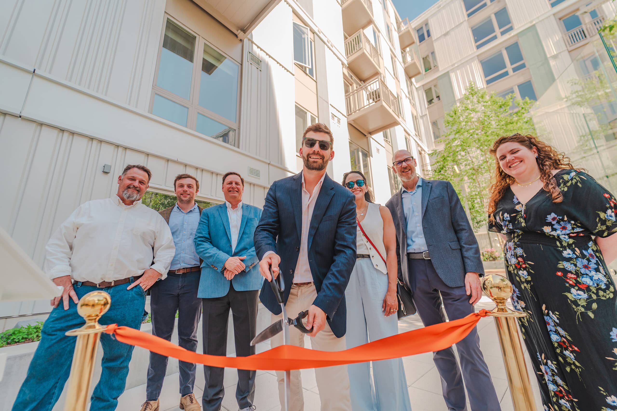 Group of people cutting an orange ribbons with large scissors in the courtyard of an apartment community