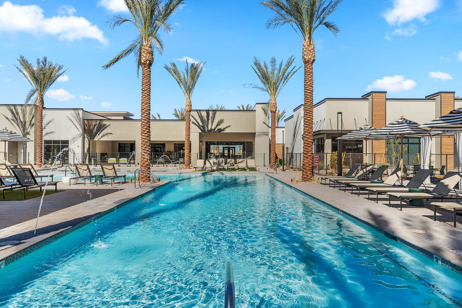 Outdoor pool at an apartment complex with fountains surrounded by palm trees and lounge chairs with striped umbrellas.