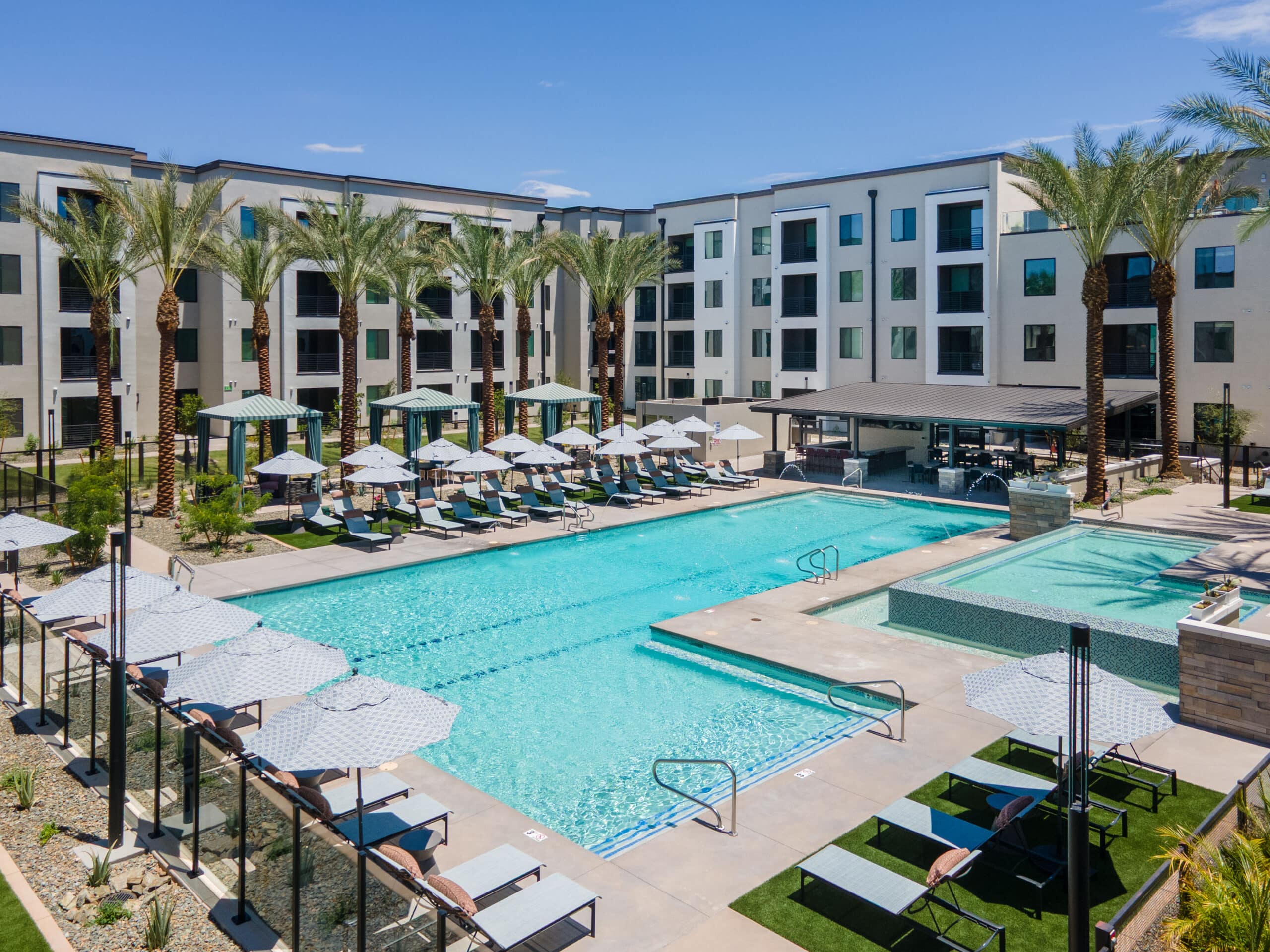 Pool at a mid-rise apartment community surrounded by lounge chairs, umbrellas, and palm trees.