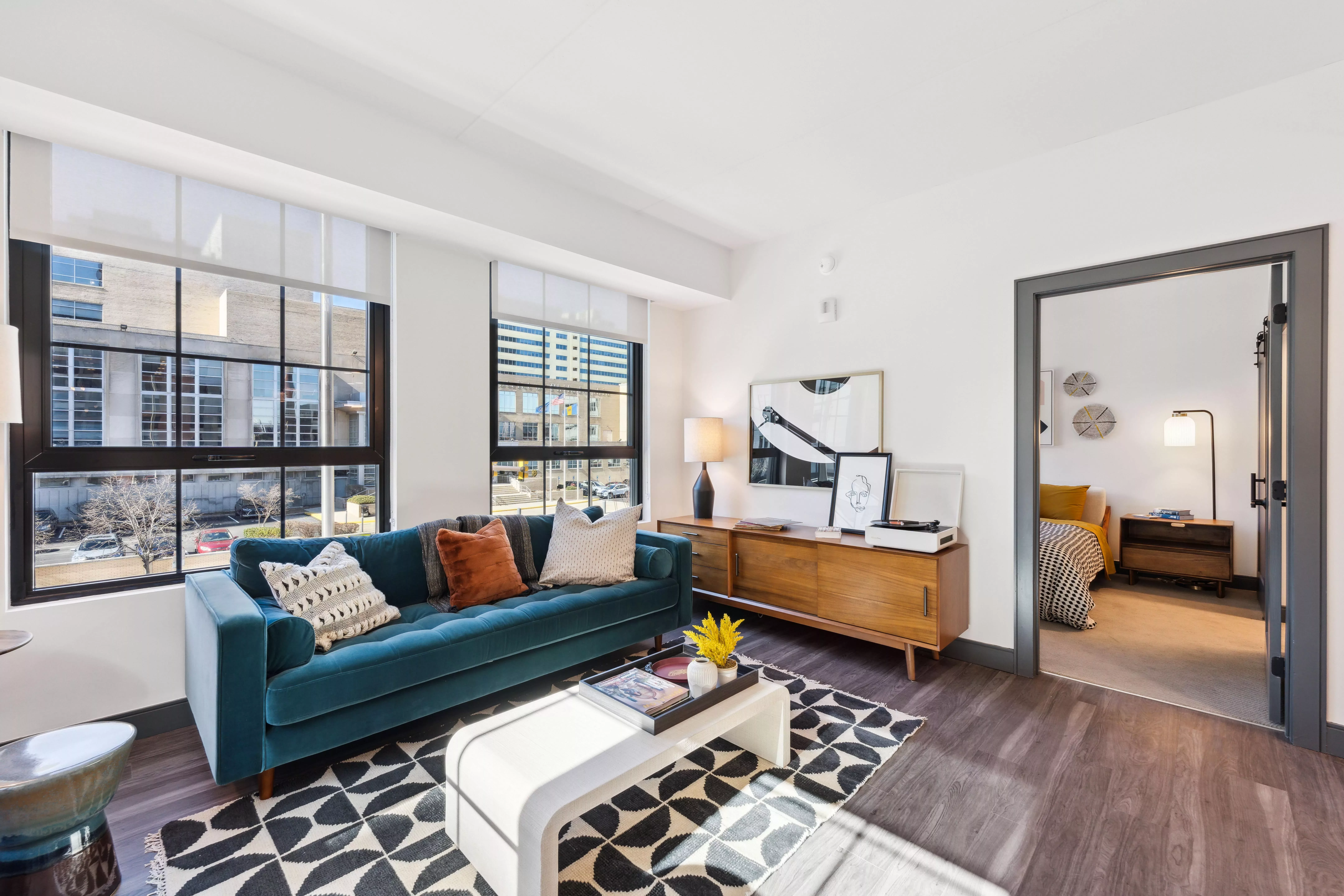 Living room with large, black-framed windows, dark hardwood-style floors, and velvet couch with the door open to the bedroom