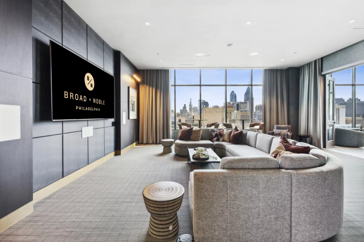 Rooftop lounge with gray couch with colorful pillows, large TV screen, and large windows with city views