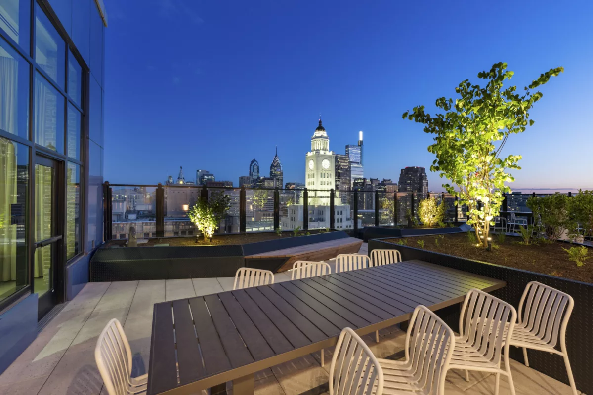 Rooftop deck at night with long table and chairs, trees with lighting, and city skyline views