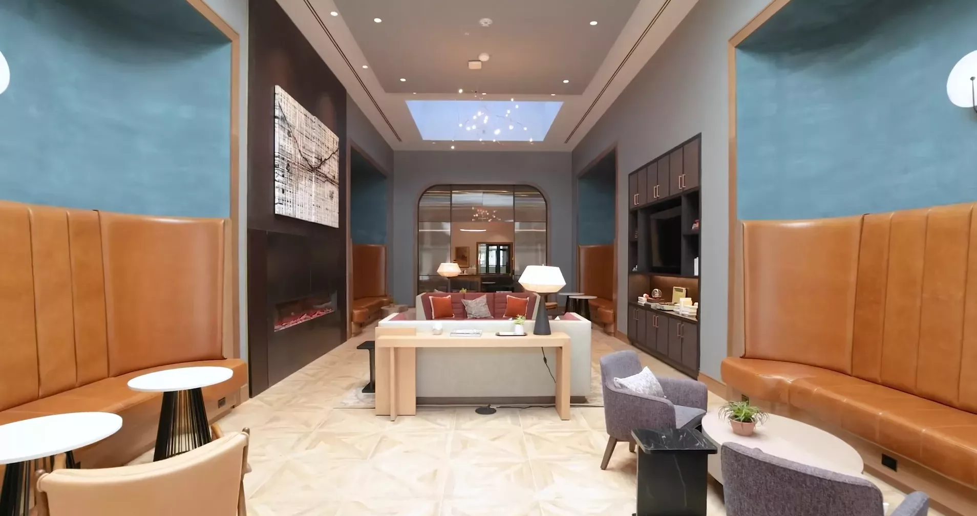 Modern lounge area with high ceilings, leather seating, tables, a large central desk, a skylight, and decorative lighting fixtures.